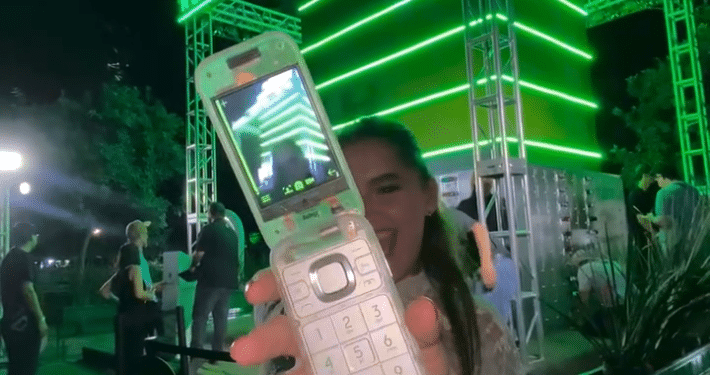Stand de Heineken de fondo con una asistent a evento mostrando el celular de The Boring Phone